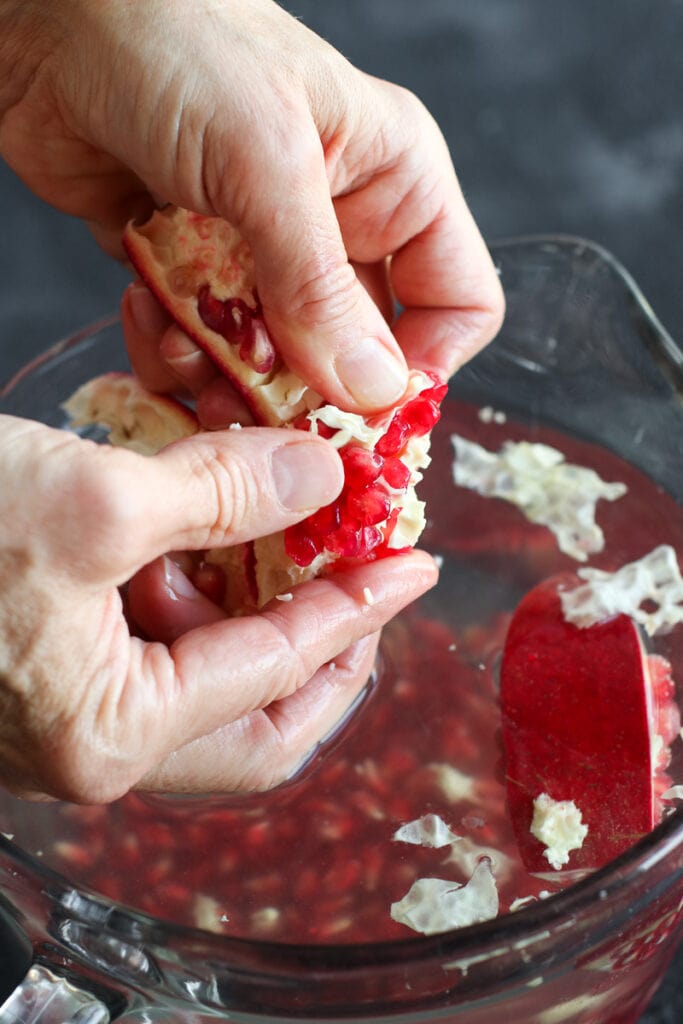 Pomegranate section in a bowl of water, two hands working to remove pomegranate arils from the membranes.