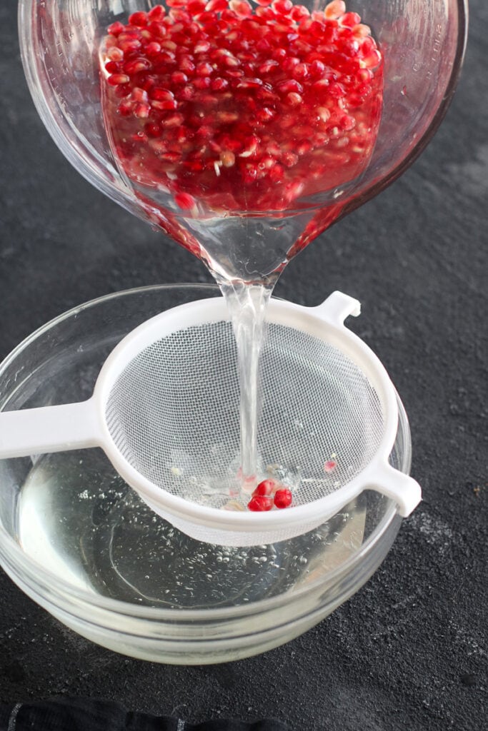 Pomegranate arils being strained from a large measuring cup of water through a white strainer 
