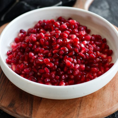 A white bowl filled with pomegranate arils, sitting on a round wooden cutting board.