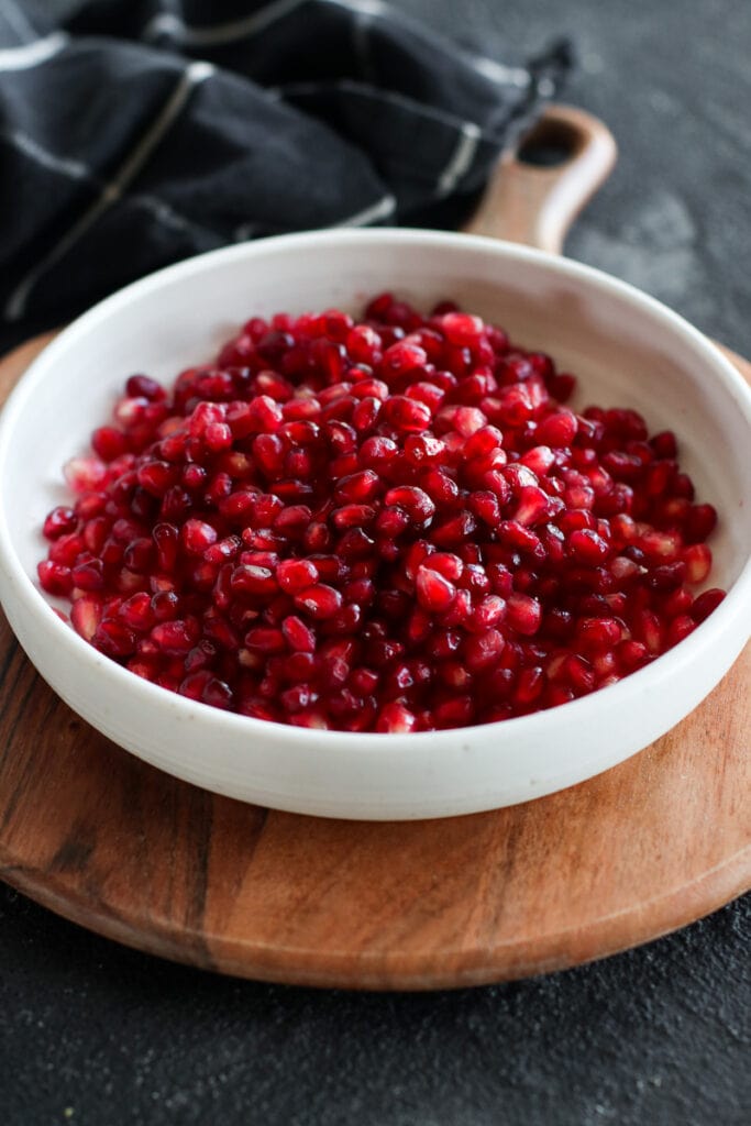 White bowl filled with pomegranate arils, on a round wooden tray.