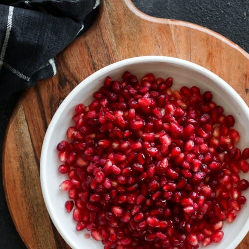 Overhead view white bowl filled with pomegranate arils on a round wooden cutting board.