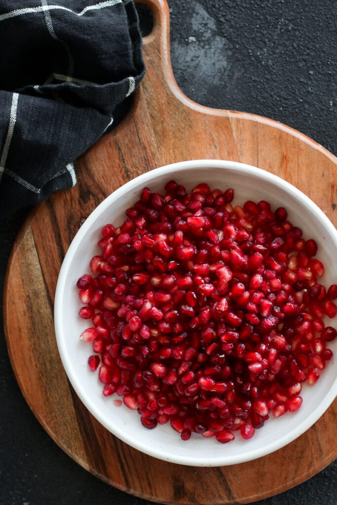 A white bowl filled with pomegranate arils on a wooden cutting board with a handle