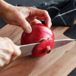 A fresh pomegranate being cut on a wooden cutting board.