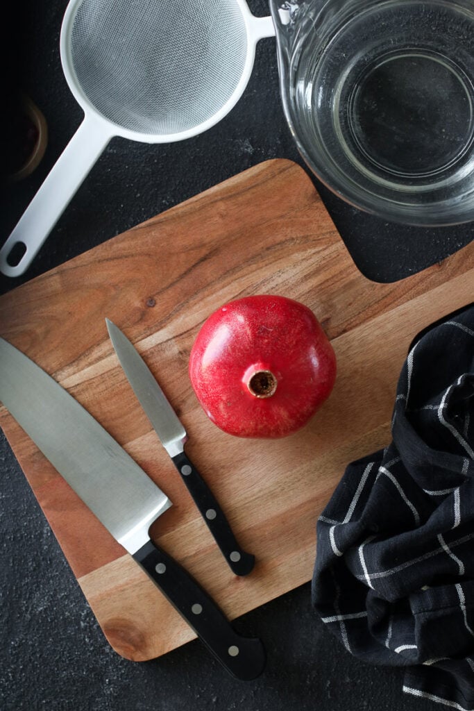A fresh pomegranate, wooden cutting board, sharp knives, a bowl of water, and white colander arranged together.