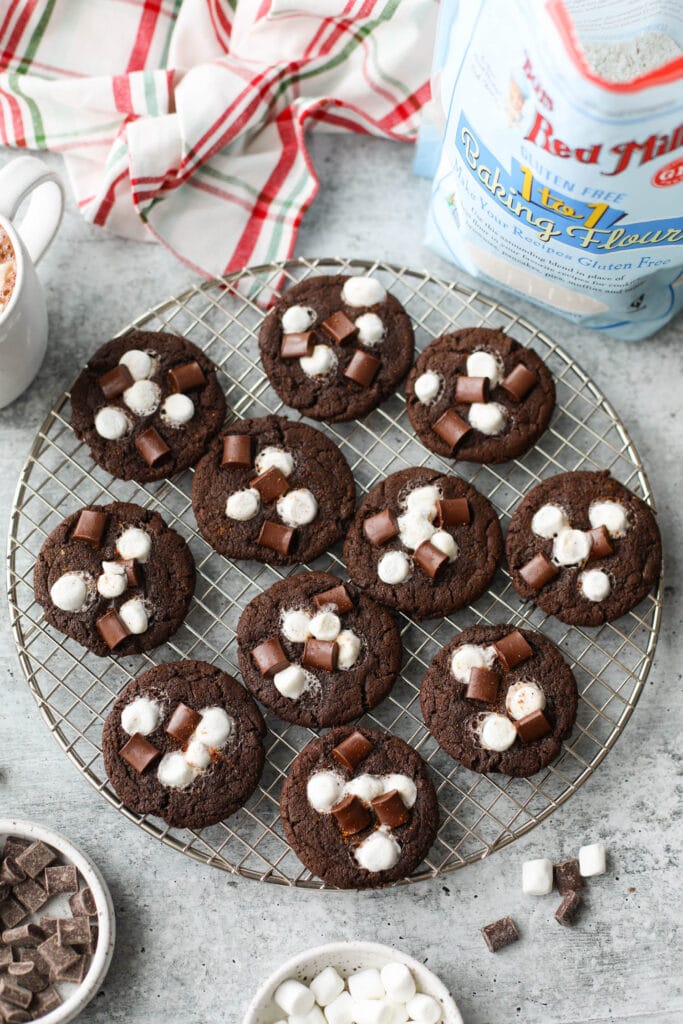 Overhead view Mexican hot chocolate cookies topped with marshmallows and chocolate chunks on wire cooling rack, bag of Bob's Red Mill Baking Flour in background.
