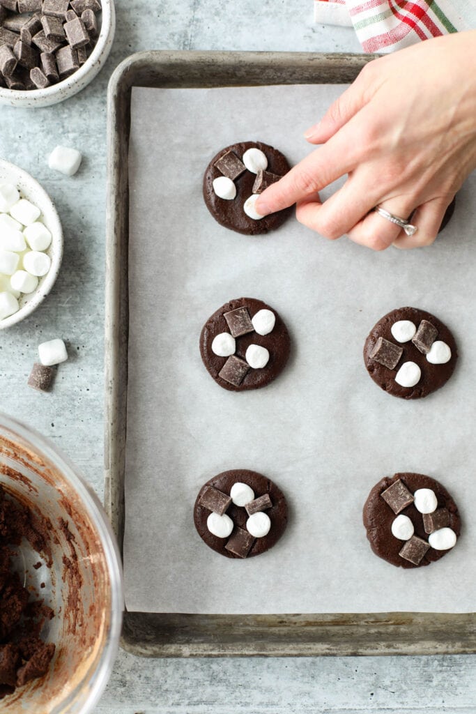 A hand pressing mini marshmallows and chocolate chunks into Mexican hot chocolate cookie sough rounds on a parchment covered baking sheet.