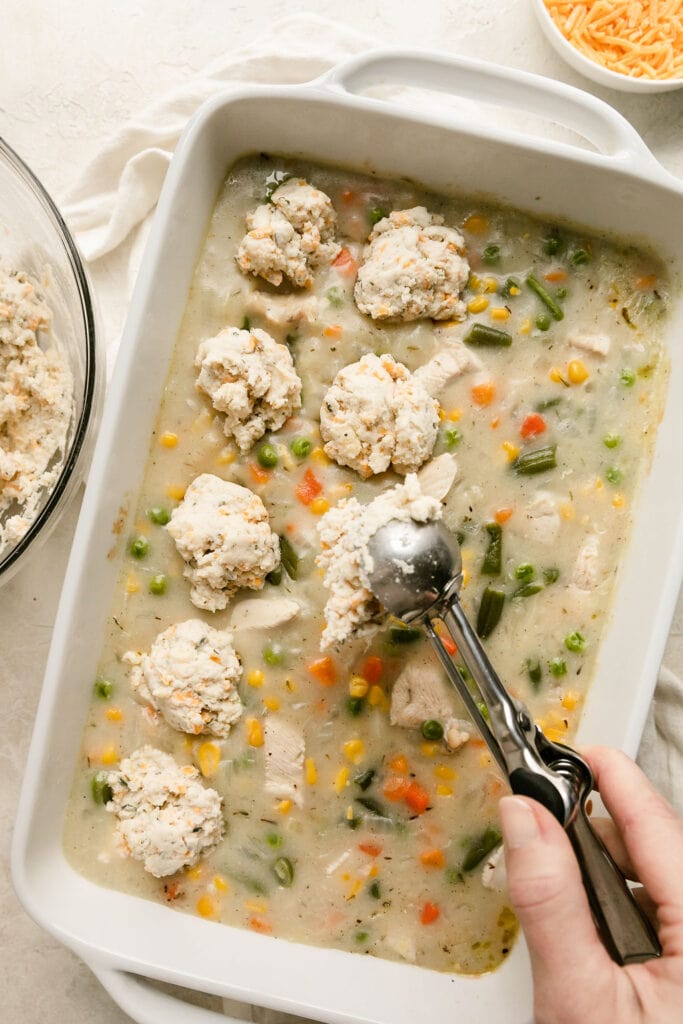 Silver scooper placing drop biscuit dough over chicken pot pie casserole in white baking dish.