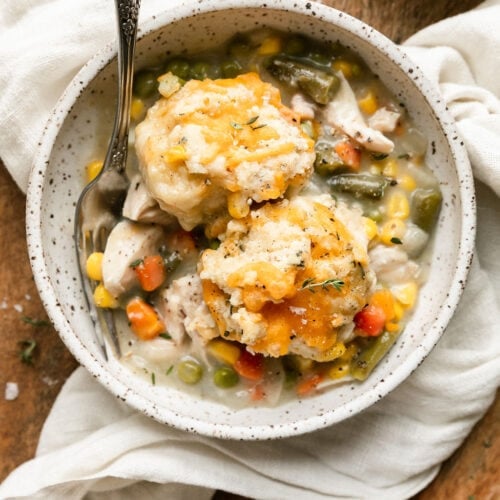 Overhead view chicken pot pie casserole serving in stone bowl on wooden cutting board, topped with cheddar drop biscuit.