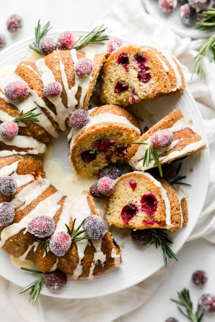 A cranberry orange olive oil bundt cake topped with white glaze, sugared cranberries and sprigs or rosemary.