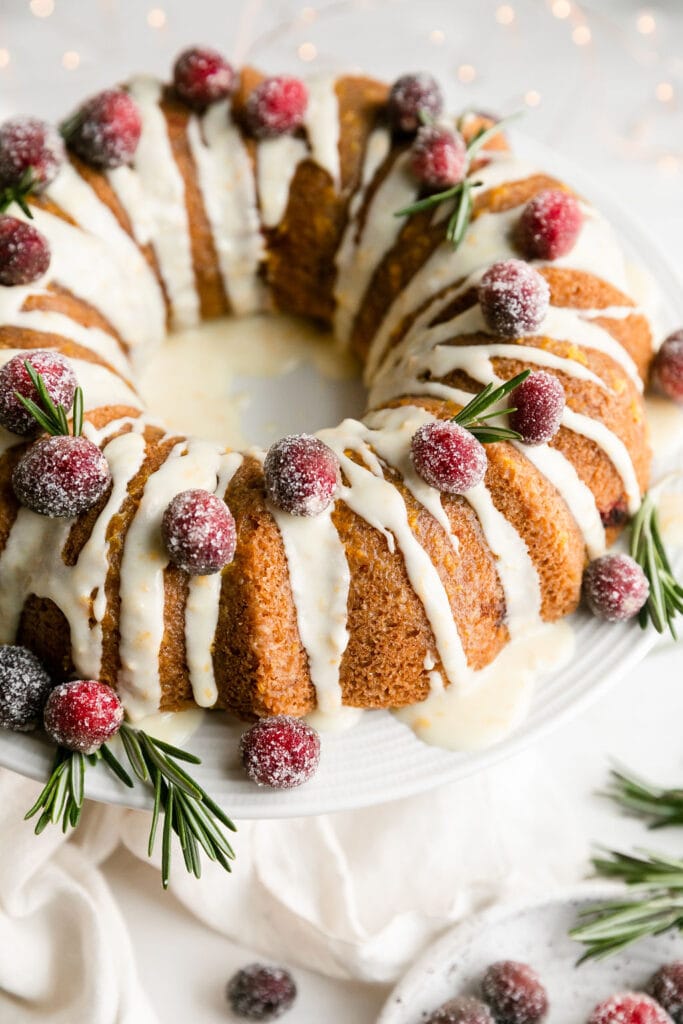 Olive oil bundt cake with cranberries and orange on white platter with white glaze.