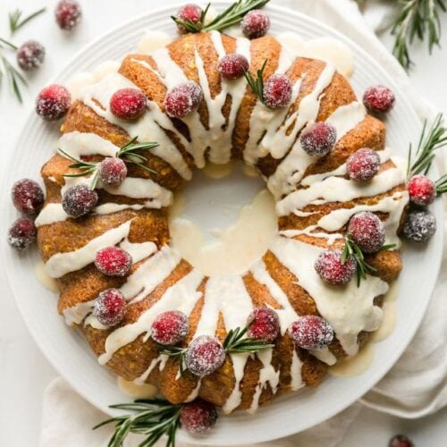 Overehad view cranberry orange olive oil bundt cake on white platter topped with icing, sugared cranberries, and rosemary sprigs.