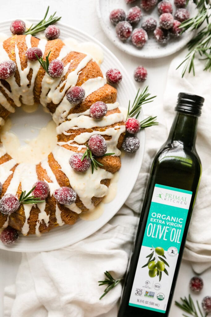 Cranberry orange olive oil bundt cake on white platter topped with white glaze, sugared cranberries and rosemary sprigs. 