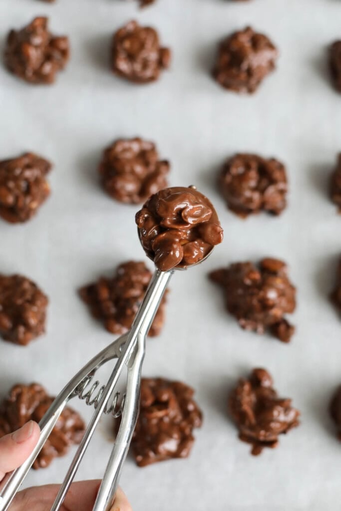 A cookie scoop filled with crockpot peanut cluster mixture over clusters lined up on parchment paper.