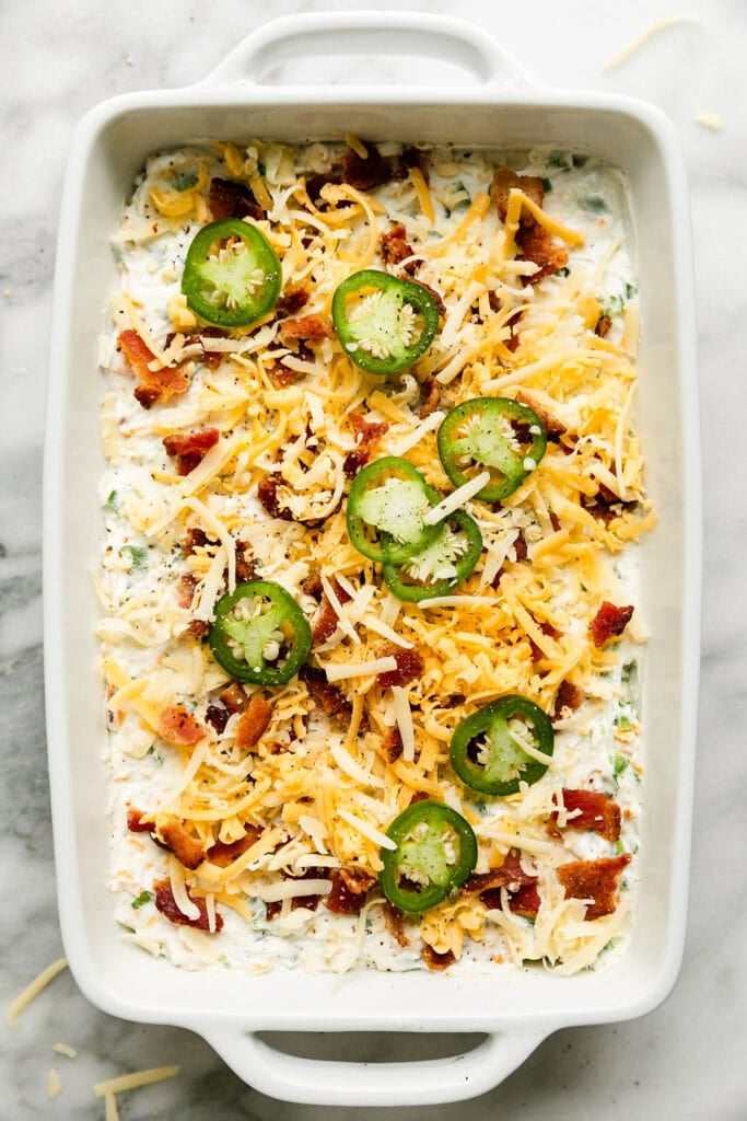 Jalapeño Popper Dip in white baking dish topped with shredded cheese, crisp bacon, and jalapeño slices ready to be baked.