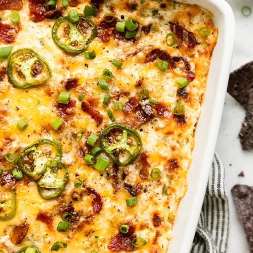 Jalapeño Popper Dip in white baking dish topped with melted cheese, jalapeño slices, and bacon pieces in a white baking dish.