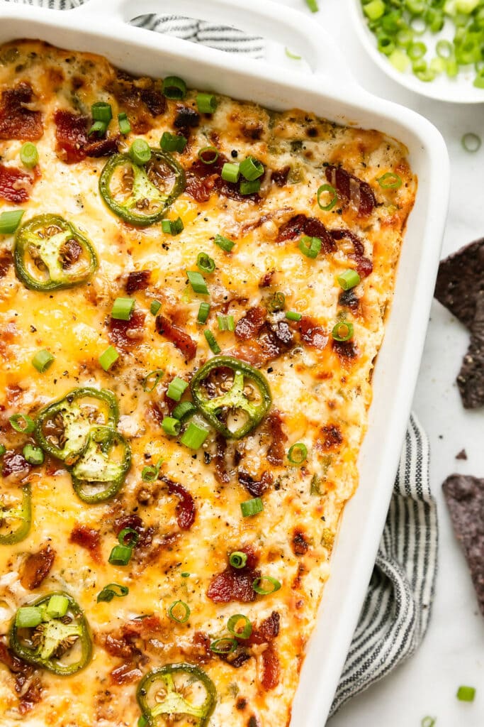 Close up view corner of white baking dish filled with jalapeño popper dip topped with melted cheese and bacon pieces. 