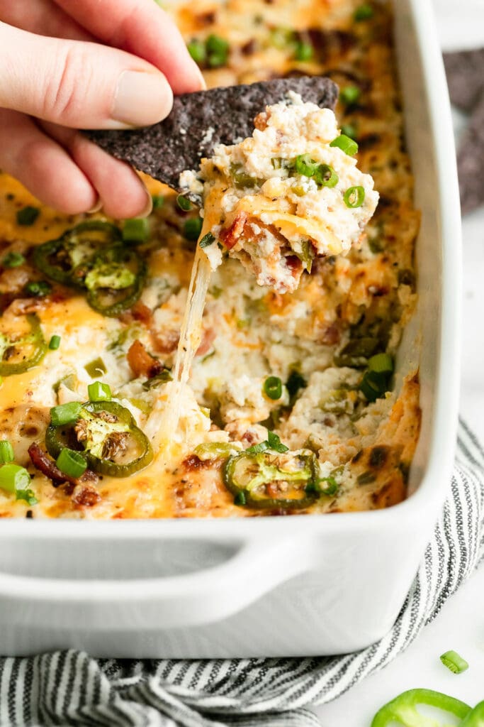 A blue corn chip scooping up jalapeño popper dip with bacon and chives from a white baking dish.