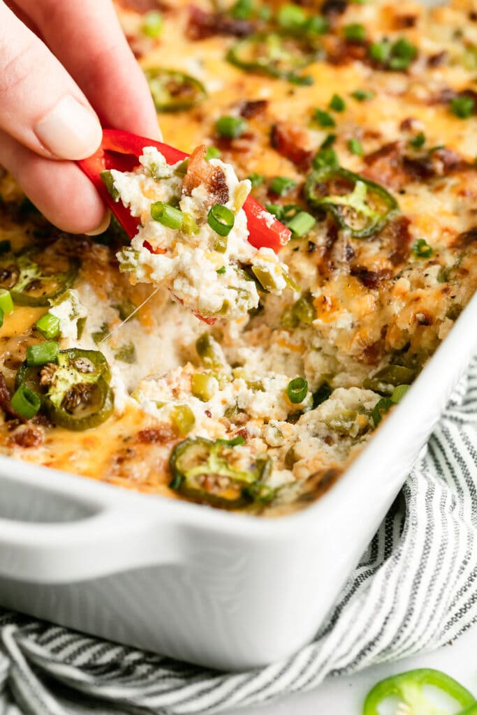 A sliced red pepper scooping up jalapeño popper dip from baking dish.