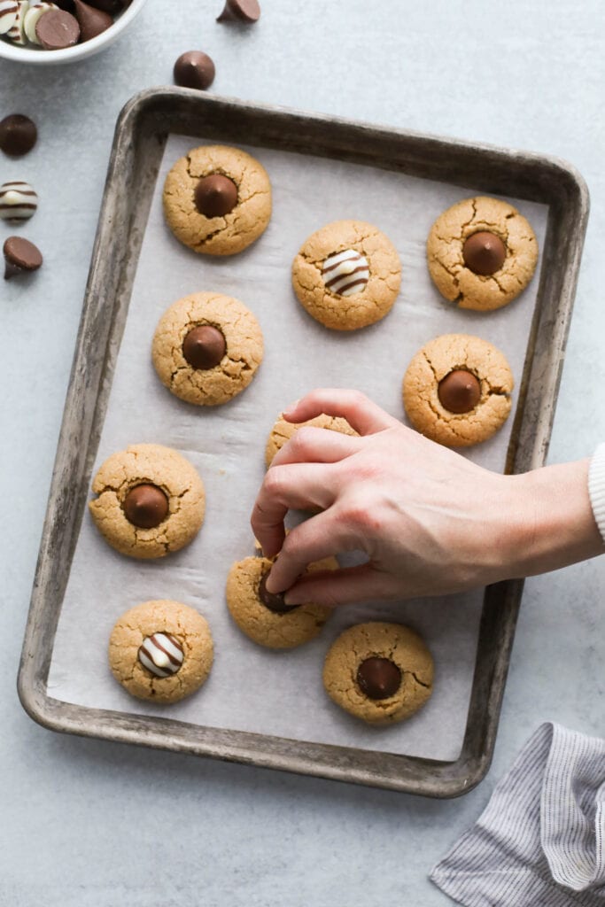 Baking sheet with peanut butter blossoms on parchment paper, chocolate kisses being pressed into baked cookies.
