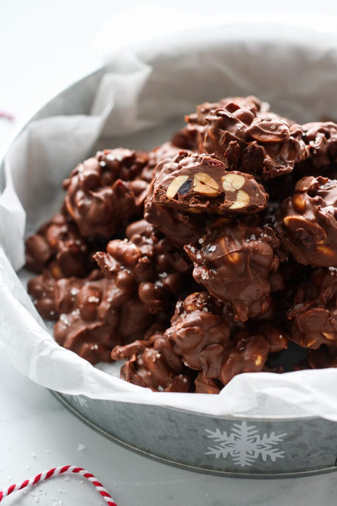 Crockpot peanut butter chocolate clusters in parchment lined Christmas cookie tin, top cluster cut in half.