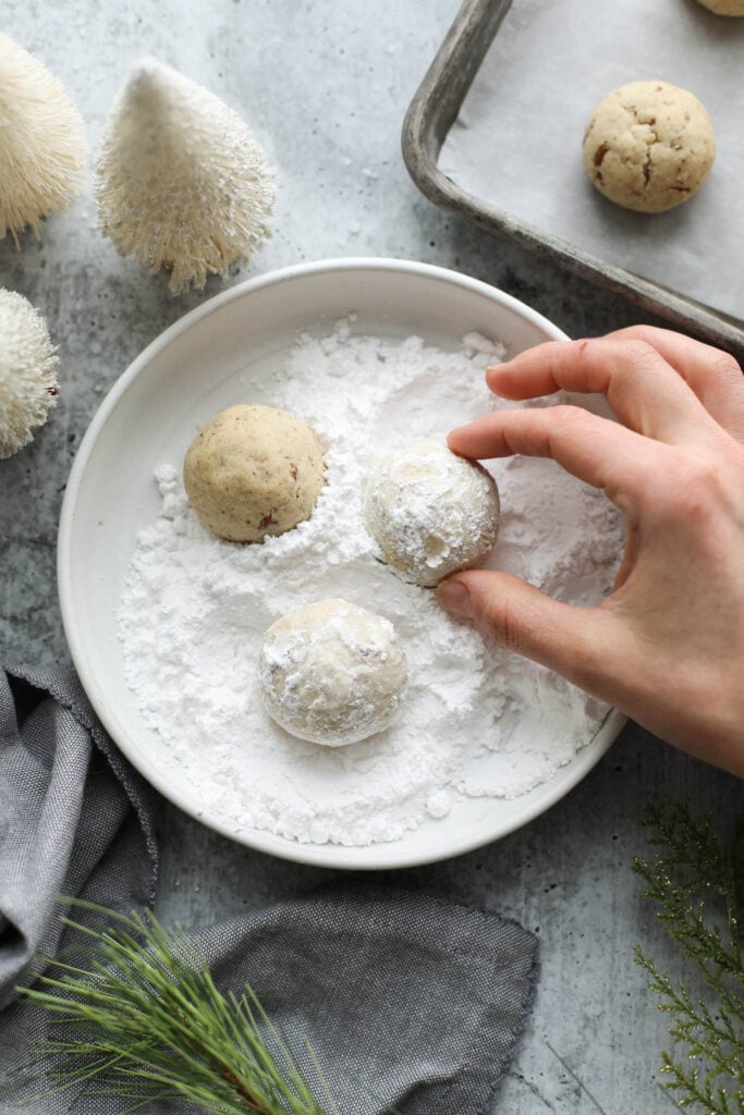 Shallow bowl filled with powered sugar, Pecan Snowball Cookies being rolled in powdered sugar.