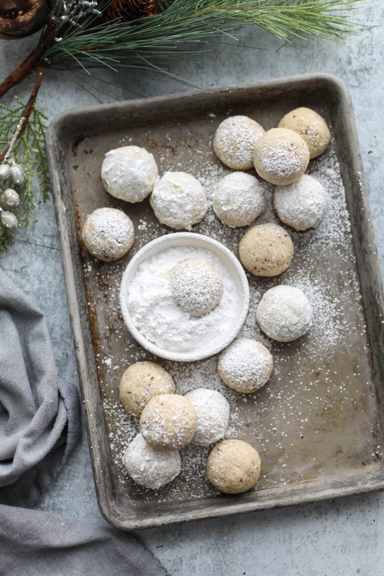 Overhead view baking sheet filled with Pecan Snowball Cookies, small bowl of powdered sugar in the center