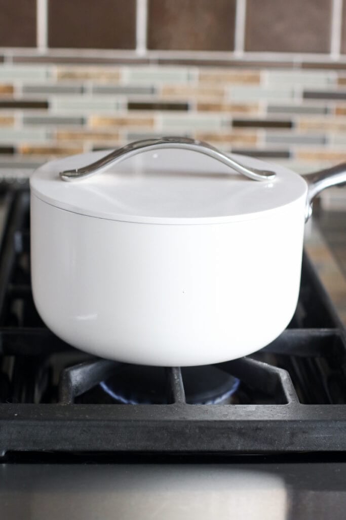 A white saucepan with lid on stovetop over open flame turned low.