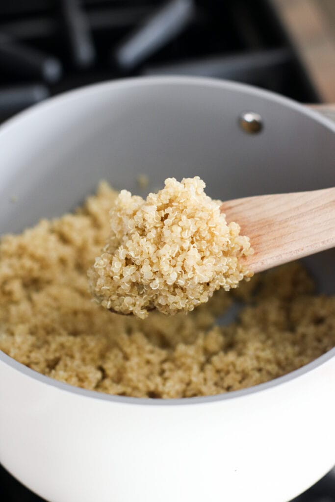 A small wooden spoon scooping up spoonful of freshly cooked quinoa from white saucepan