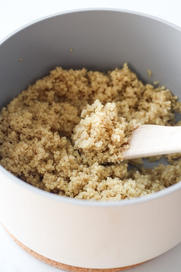A small wooden spoon scooping up spoonful of fresh cooked quinoa from white saucepan.