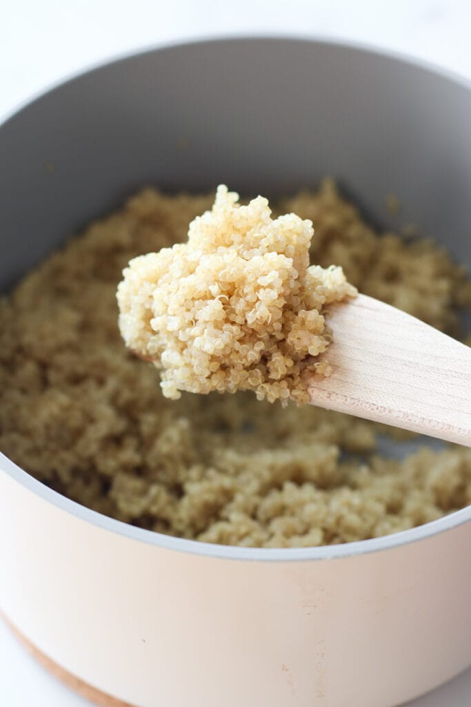 Close up view wooden spoon with scoop of cooked quinoa over a white saucepan filled with quinoa
