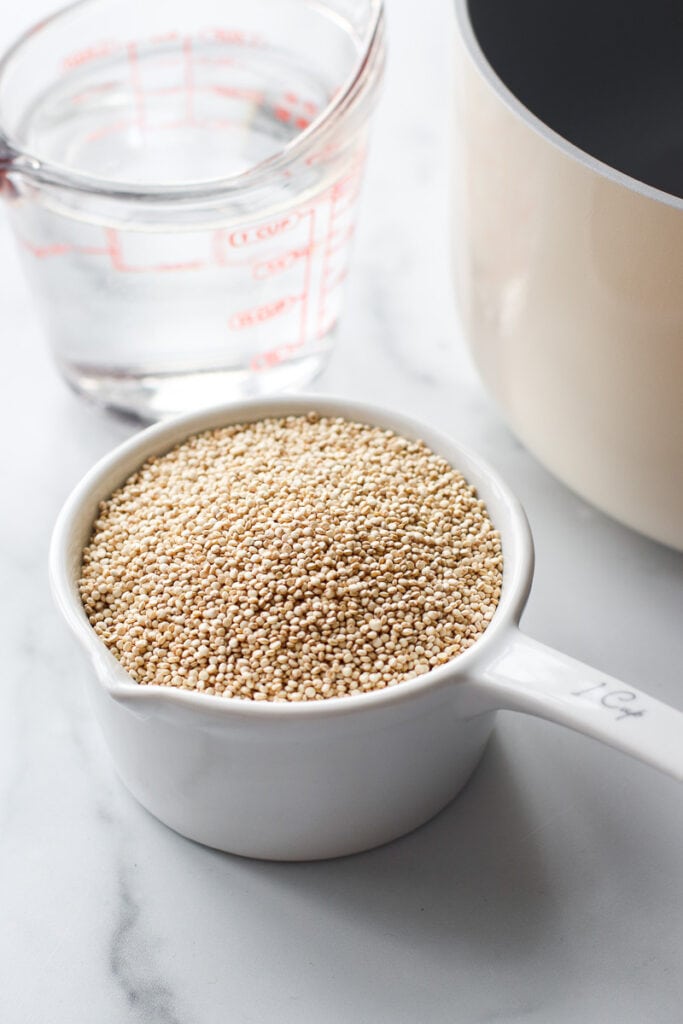 Measuring cup filled with uncooked quinoa, measuring cup with water, white saucepan to the corner