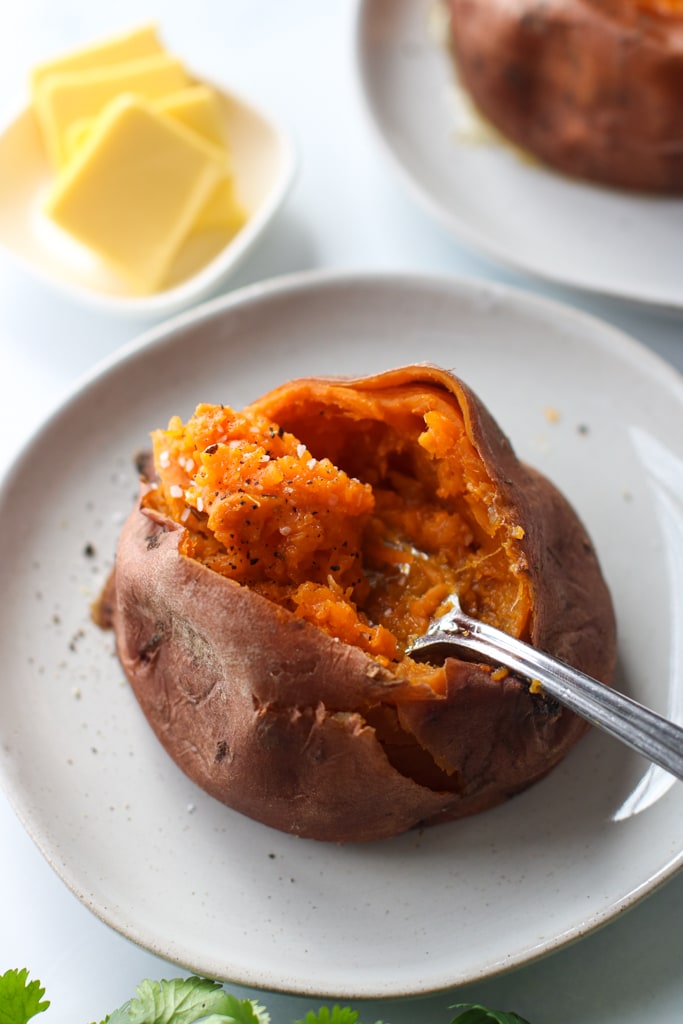 Overhead view instant pot sweet potato cut open, sprinkled with sea salt and pepper on white plate with fork lifting up fluffy orange sweet potato