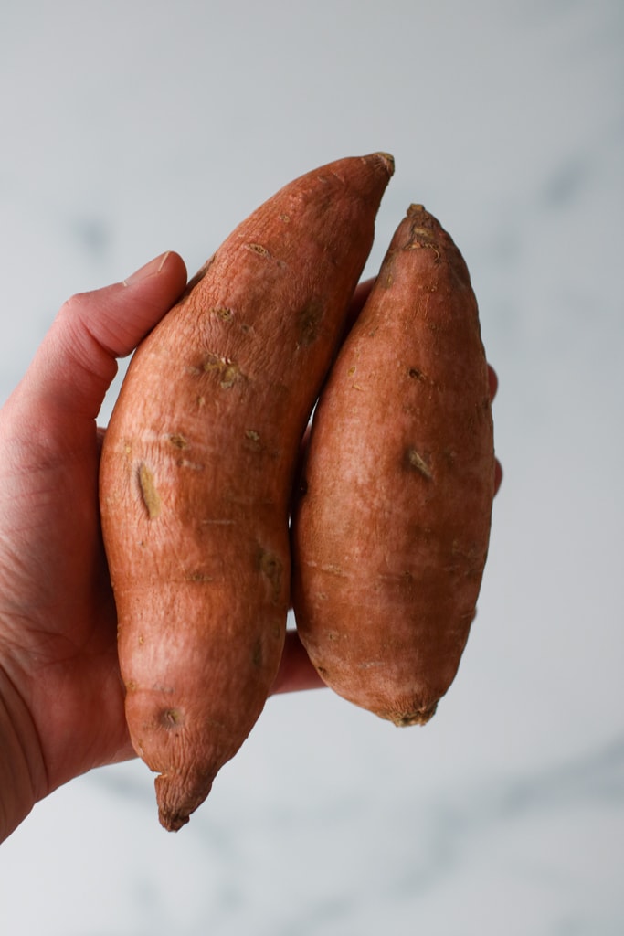 A hand holding two small sweet potatoes against marble background.