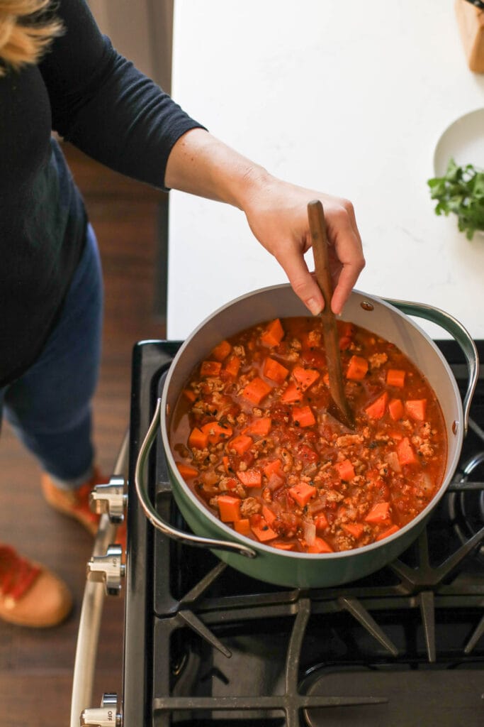 A green Caraway pot filled with sweet potato chili cooking on stovetop being stirred with wooden spoon