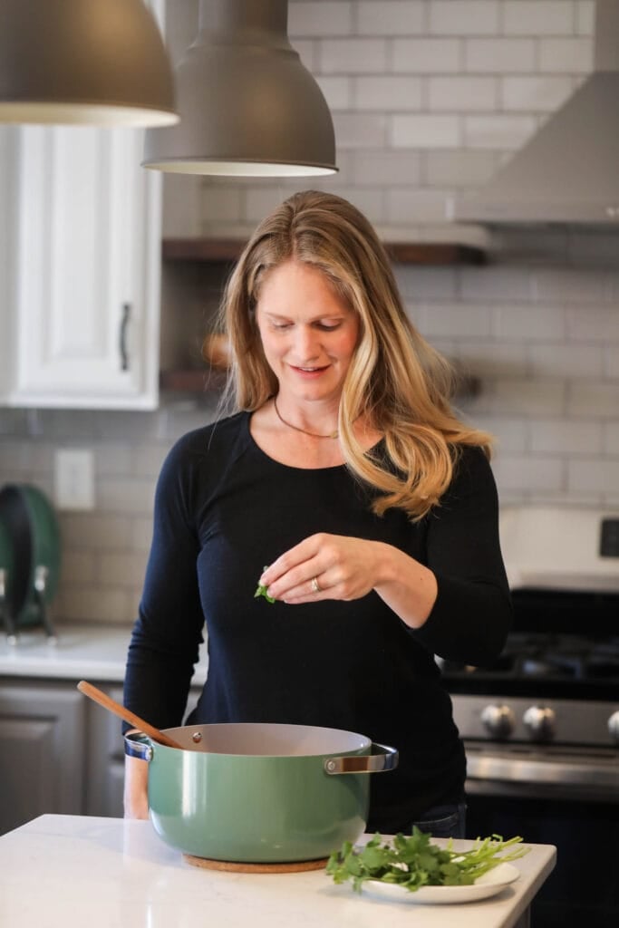 A woman standing in kitchen behind island with green Caraway pot on island, sprinkling herbs in pot