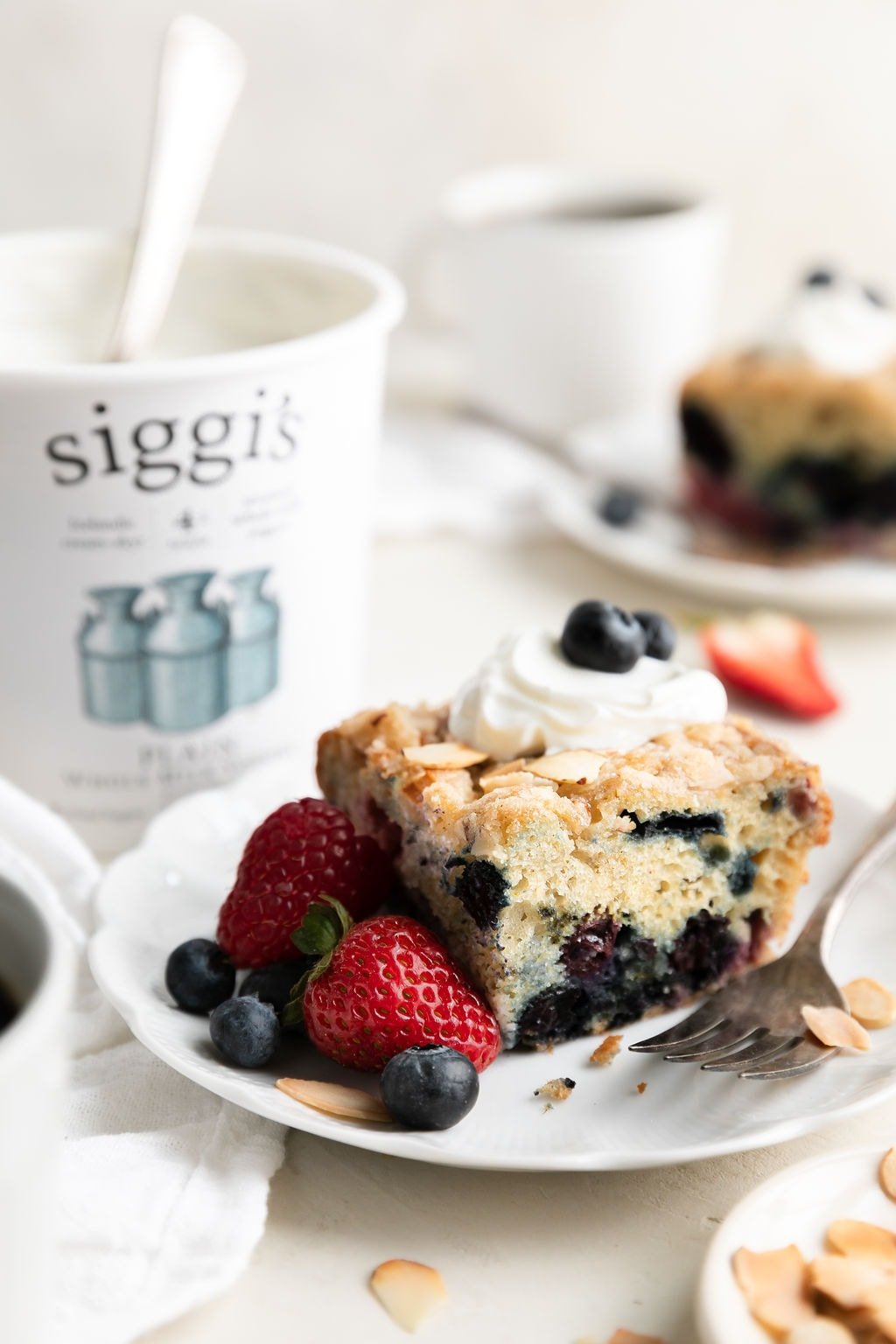 A slice of mixed berry breakfast cake on white plate topped with yogurt and fresh berries. 