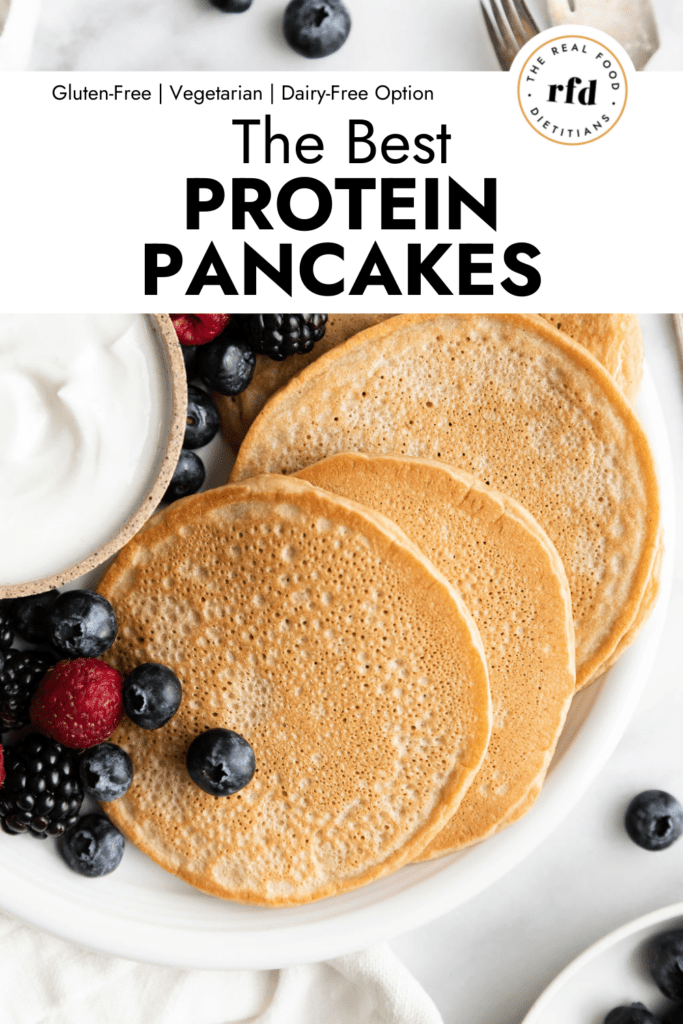 Several protein pancakes on plate with fresh berries and small bowl of yogurt on side.