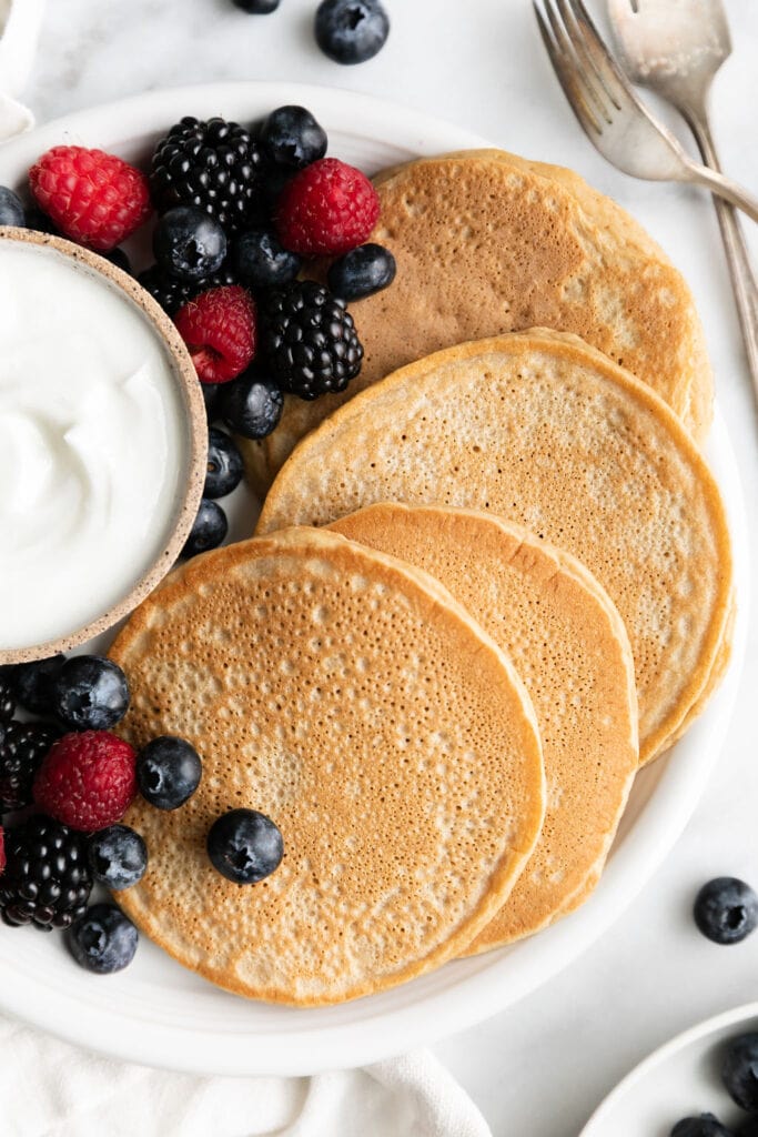 Four protein pancakes on white plate with fresh berries and small bowl of yogurt.