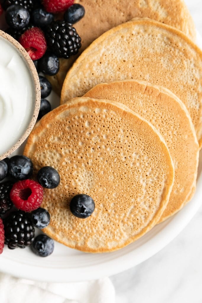 Several protein pancakes on white plate with fresh berries and bowl of yogurt.