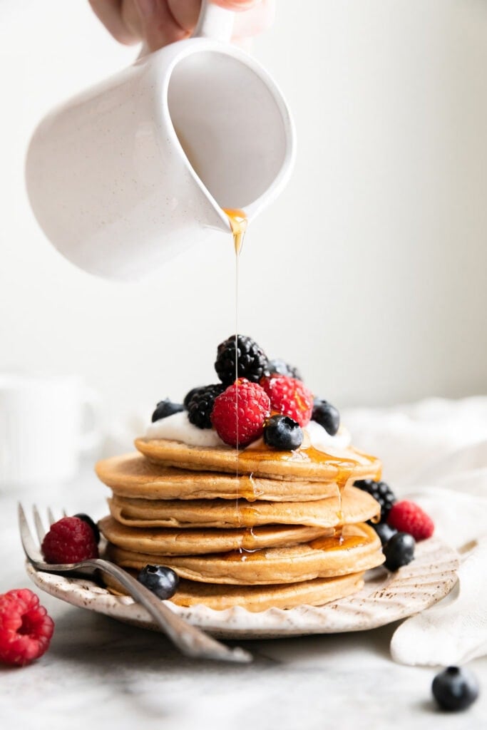 Maple syrup being drizzled over top tall stack of protein pancakes on white plate topped with fresh berries.