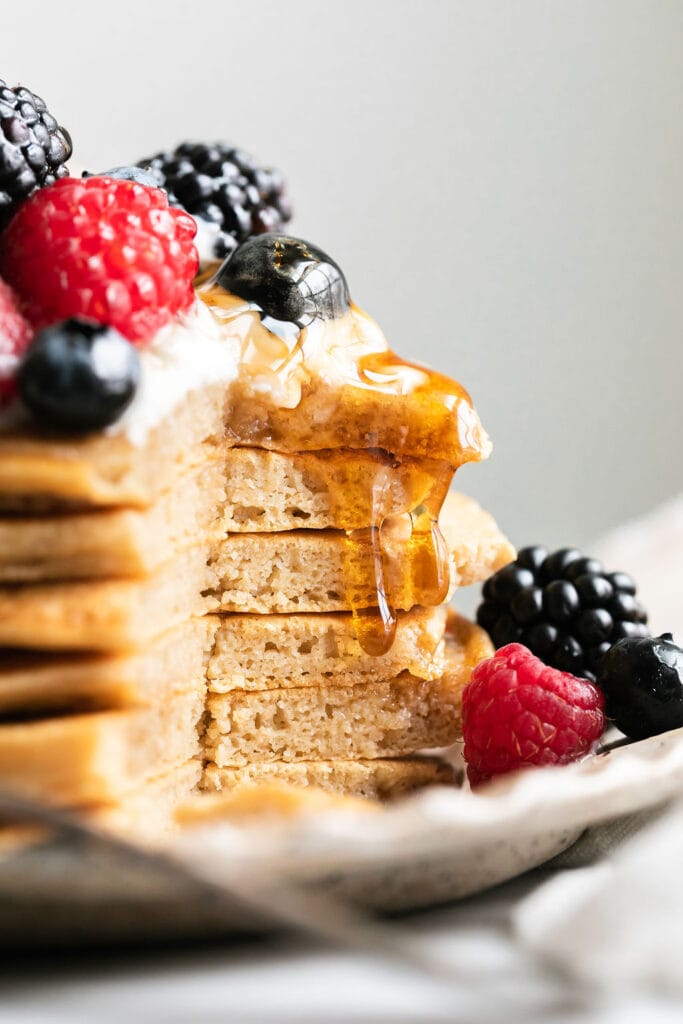 A tall stack of protein pancakes on plate, slice cut out to show inside texture of pancakes, maple syrup dripping down edge.