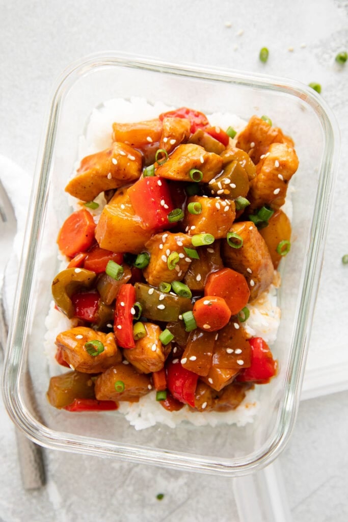 Overhead view of sweet and sour chicken with vegetables and topped with sesame seeds and green onions in glass container. 