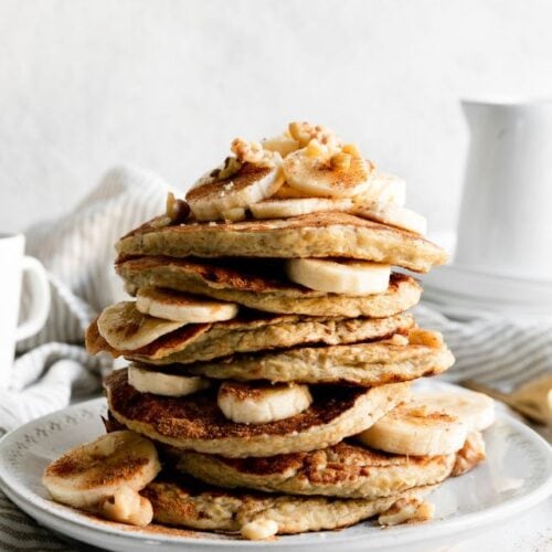 Tall stack of banana oatmeal pancakes with banana slices and walnuts between pancakes on white plate.
