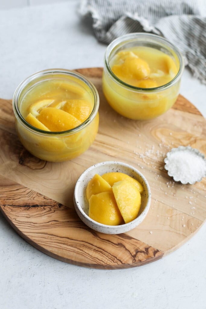 Two jars of preserved lemons, a small dish of lemon quarters and some salt on a cutting board