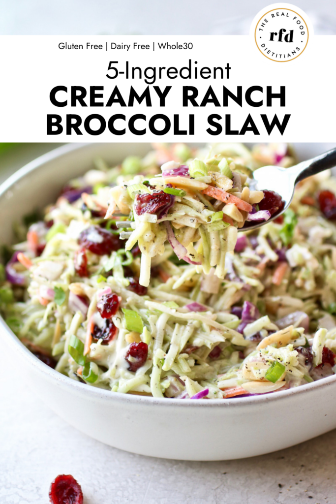 A forkful of creamy ranch broccoli slaw with dried cranberries and almonds being lifted up over a white serving bowl filled with slaw
