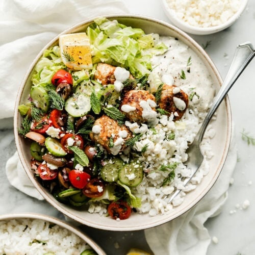 Overhead view bowl filled with Greek chicken meatballs served over greens, cucumbers, tomatoes, kalamata olives and tzatziki sauce.