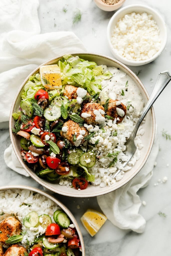 Overhead view Greek bowl of greens, rice, tomatoes, cucumbers, chicken meatballs and topped with tzatziki sauce.