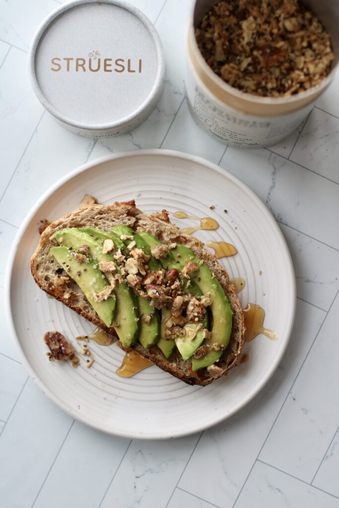 Overhead view sourdough toast topped with slices of avocado, honey, and Struesli, grain-free granola pieces.