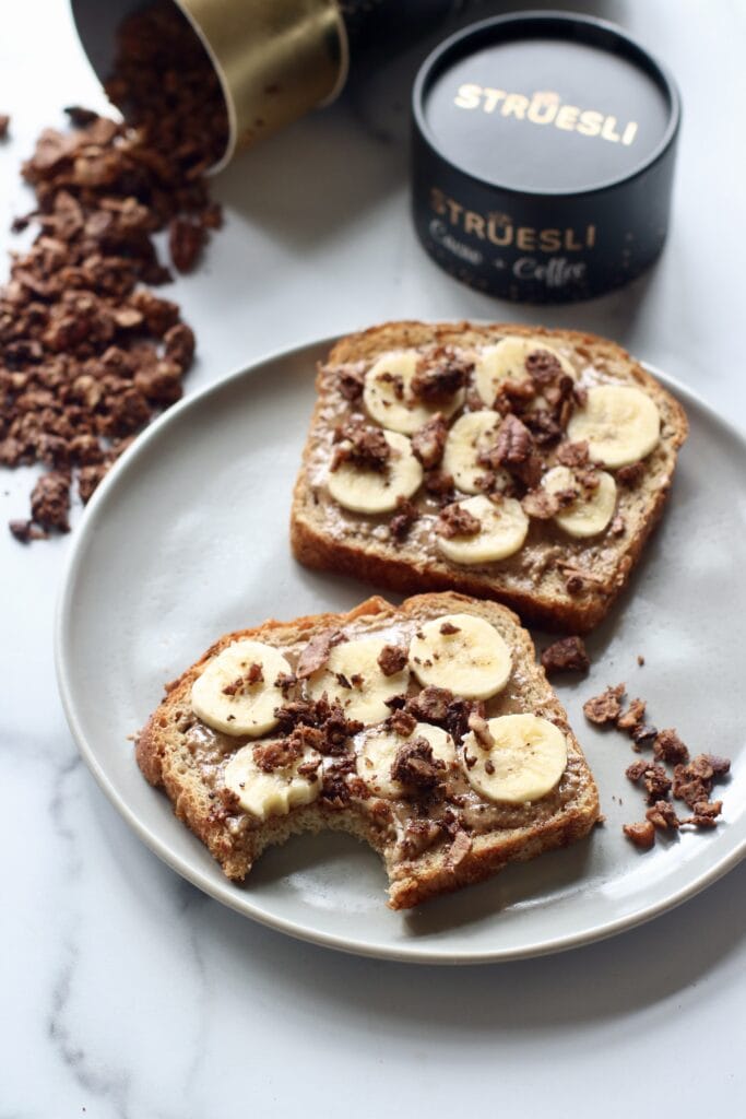 Two pieces toast with peanut butter, banana slices, and grain free granola brand Struesli, on white plate