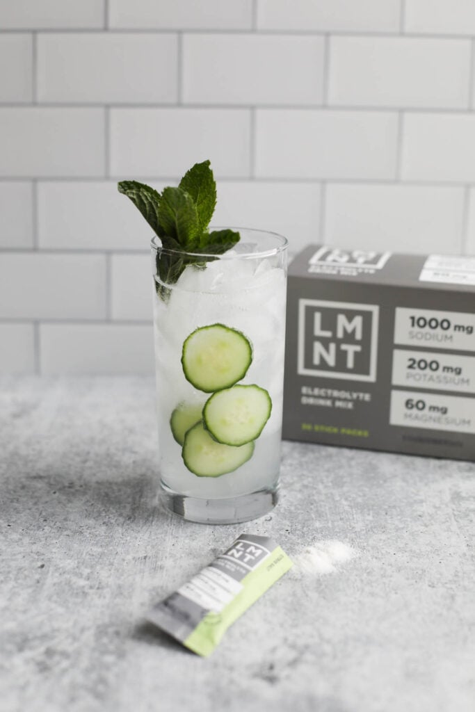 Close up view of a glass of LMNT electrolyte drink with a mint sprig and sliced cucumbers. 